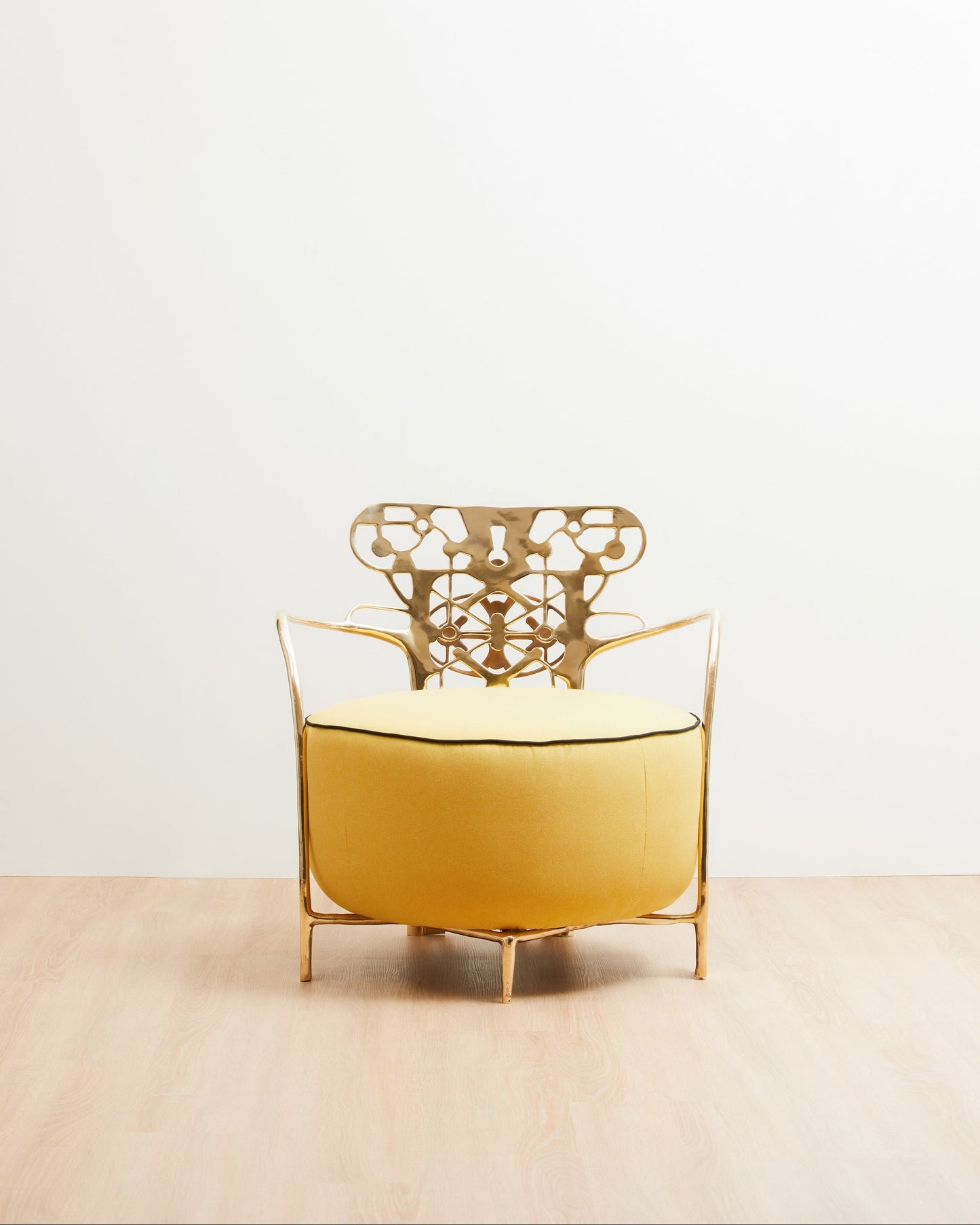A modern accent chair with a yellow upholstered seat and a gold metal frame, featuring a decorative openwork backrest, set against a minimalist white wall and light wood floor.