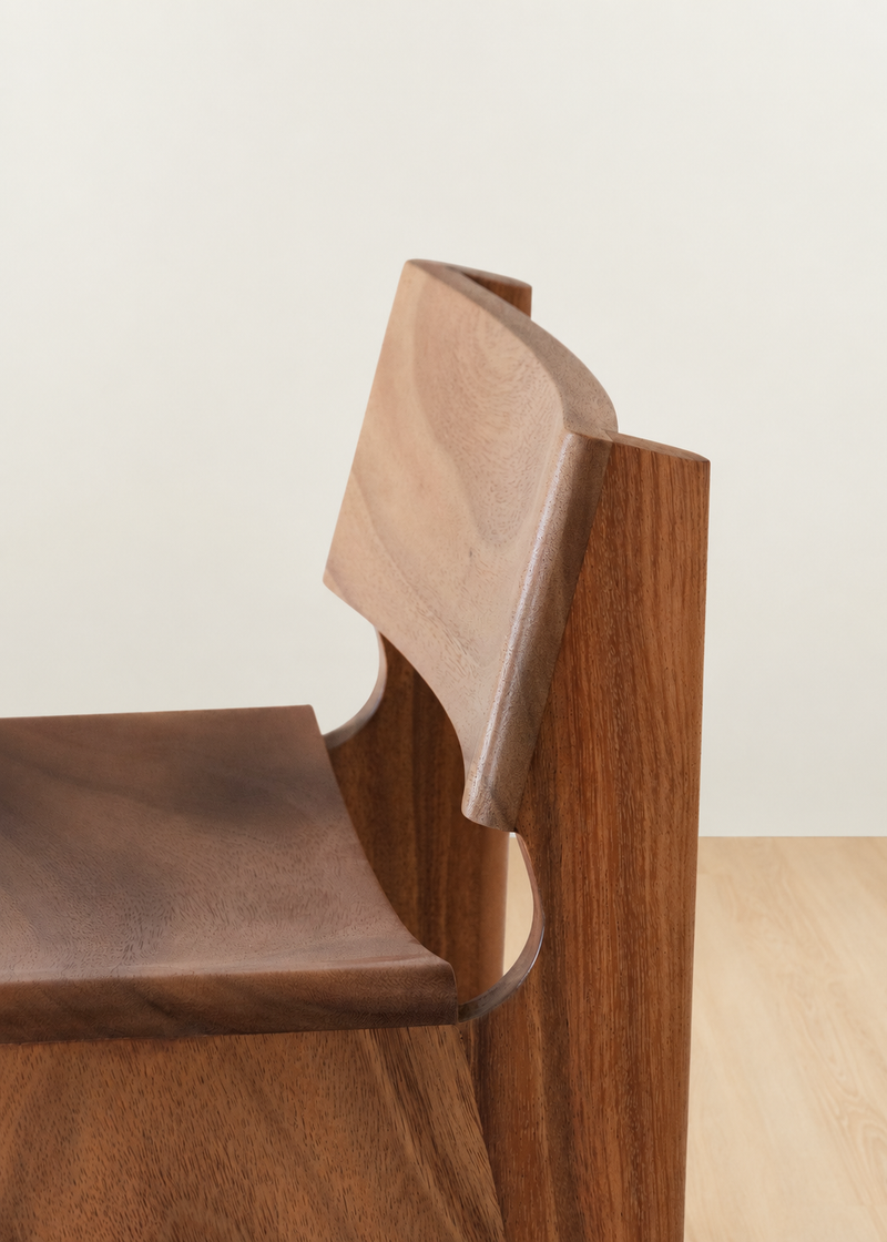 Close-up of a wooden chair showing the backrest and seat in warm medium-brown wood tones against a light neutral wall and pale floor.