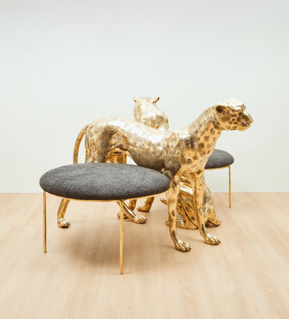 A set of two modern animal-shaped side tables or stools in polished gold-tone metal, with one round cushioned stool in front featuring a dark gray upholstered seat and two matching gold animal forms behind it, photographed in a minimalist studio setting with a light wood floor and white wall.