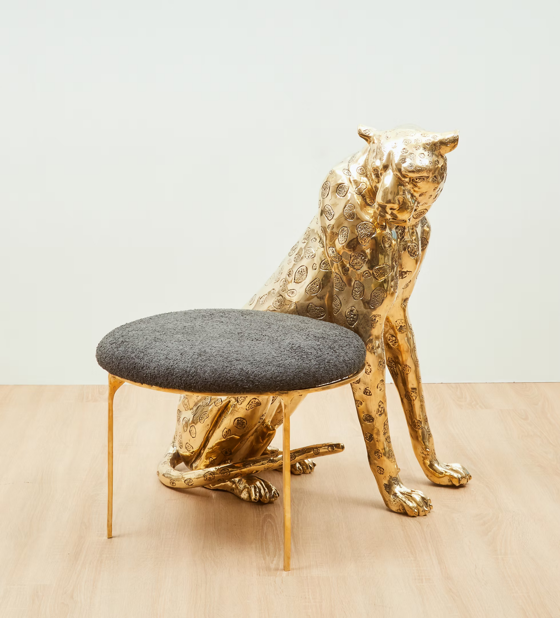 A modern gold-finished cat-shaped stool with a textured gray upholstered round seat and slim gold legs, set against a neutral light gray wall and light wood floor.