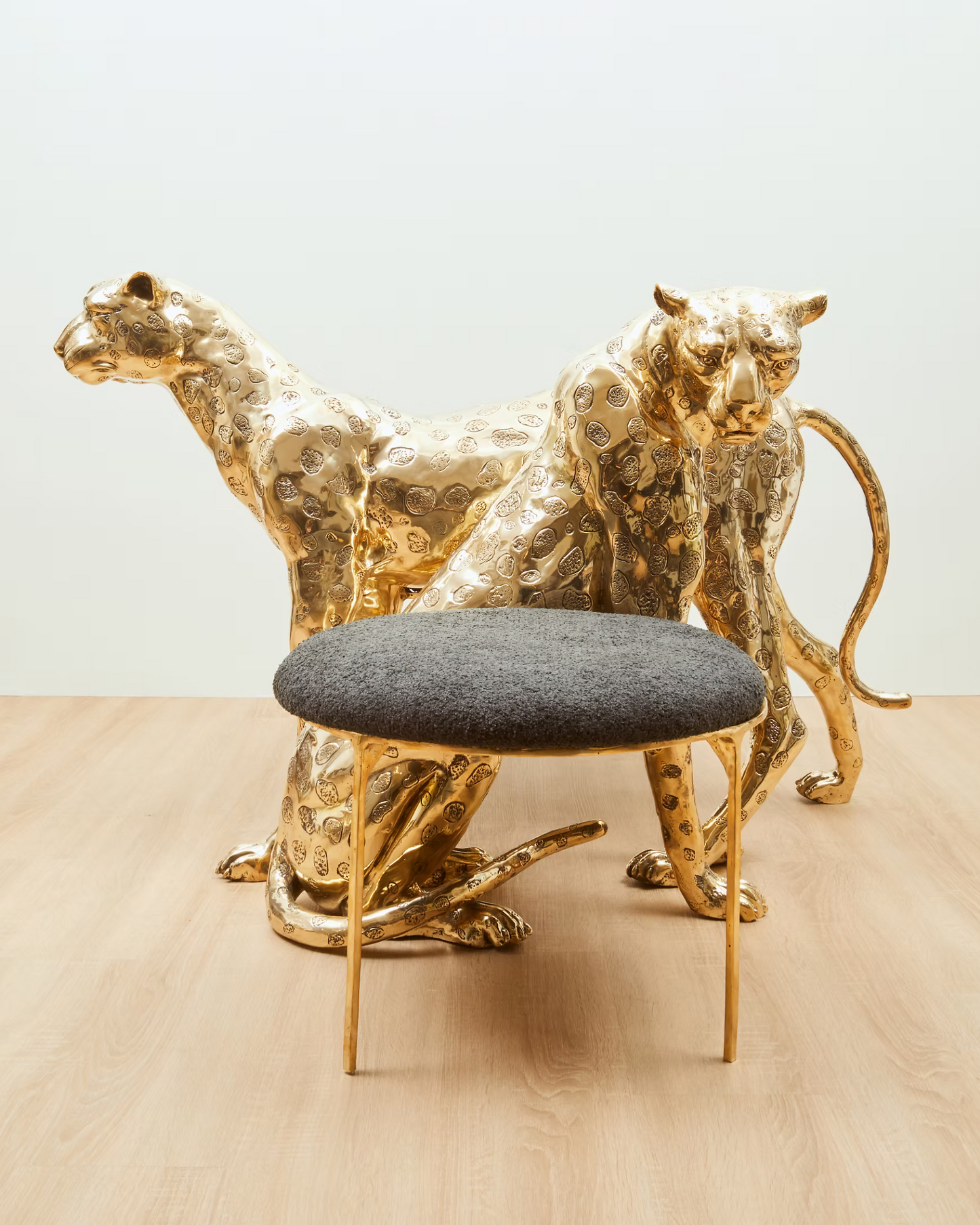A pair of ornate gold-finished leopard sculptures behind a small round stool with a dark gray upholstered seat and slim gold legs, set on a light wood floor against a plain light gray wall.