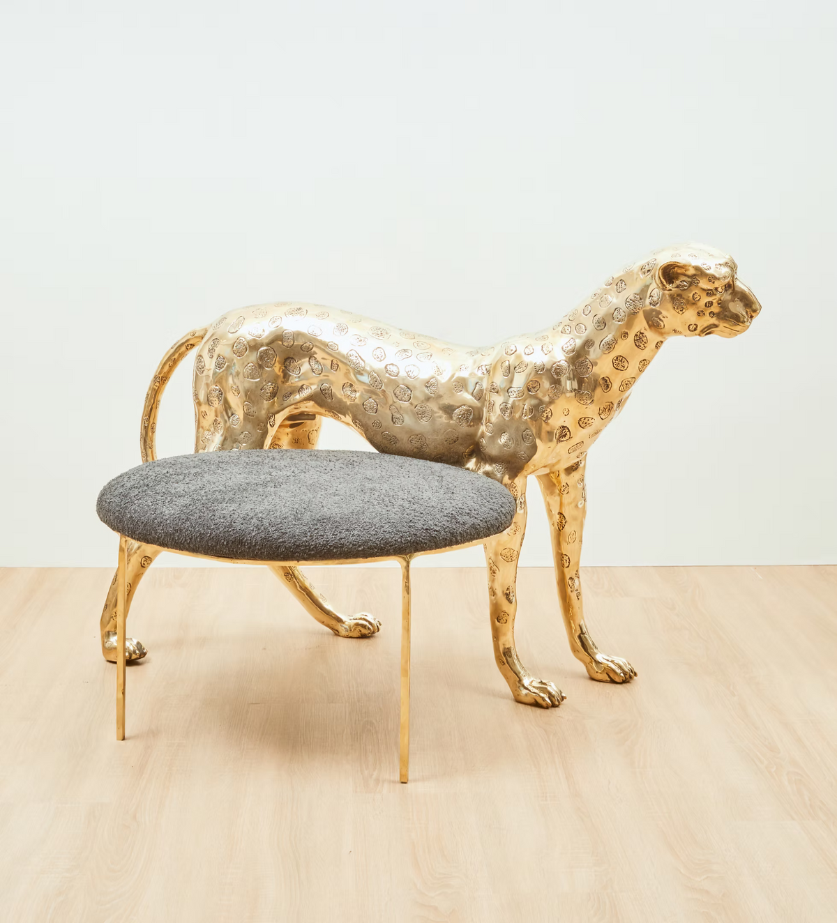 A modern leopard-shaped metallic gold sculpture paired with a round gray upholstered ottoman on slim gold legs, set against a minimalist white wall and light wood floor.