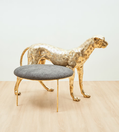 A modern leopard-shaped metallic gold sculpture paired with a round gray upholstered ottoman on slim gold legs, set against a minimalist white wall and light wood floor.