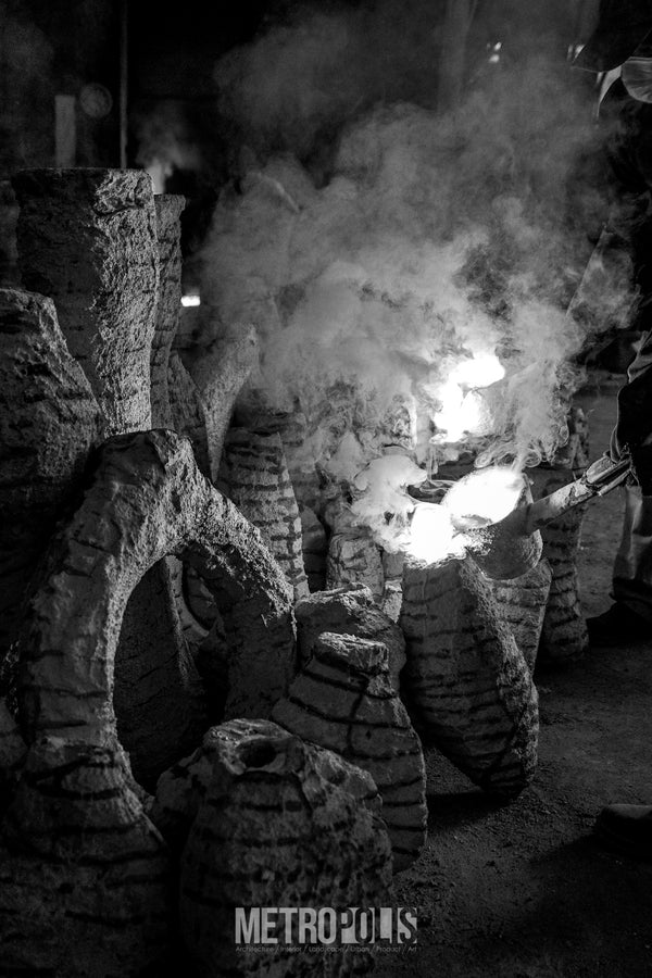 Black and white photograph of a textured stone or clay vessel arrangement with bright glowing embers or fire in the center, surrounded by smoke and dark surroundings. 