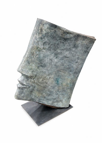 A bronze textured, sculptural book-shaped object with a metallic finish in muted blue-gray tones, displayed on a dark rectangular base against a clean white background.