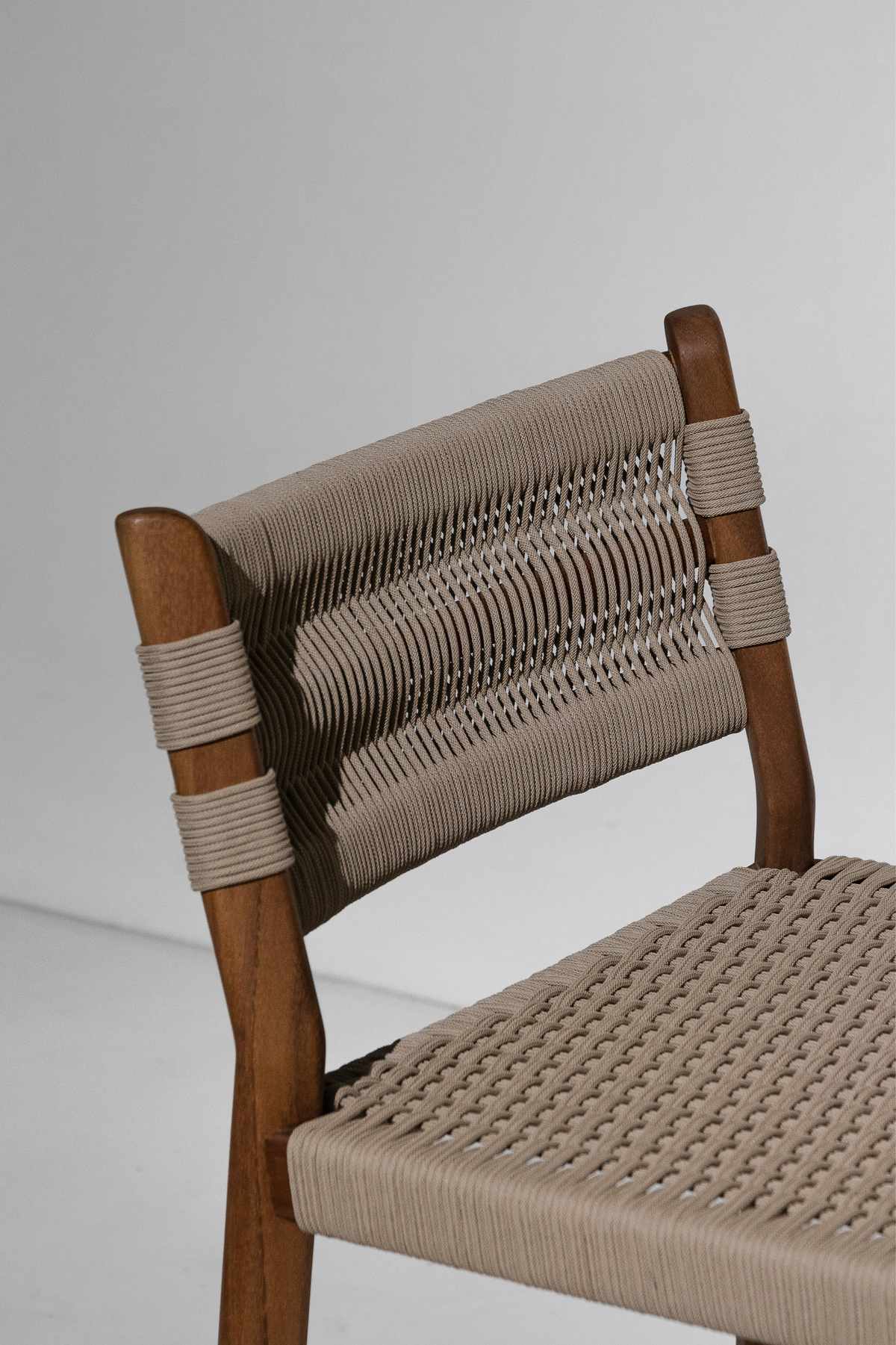 A close-up of a wooden dining chair with a woven beige rope seat and backrest, set against a plain light gray background.
