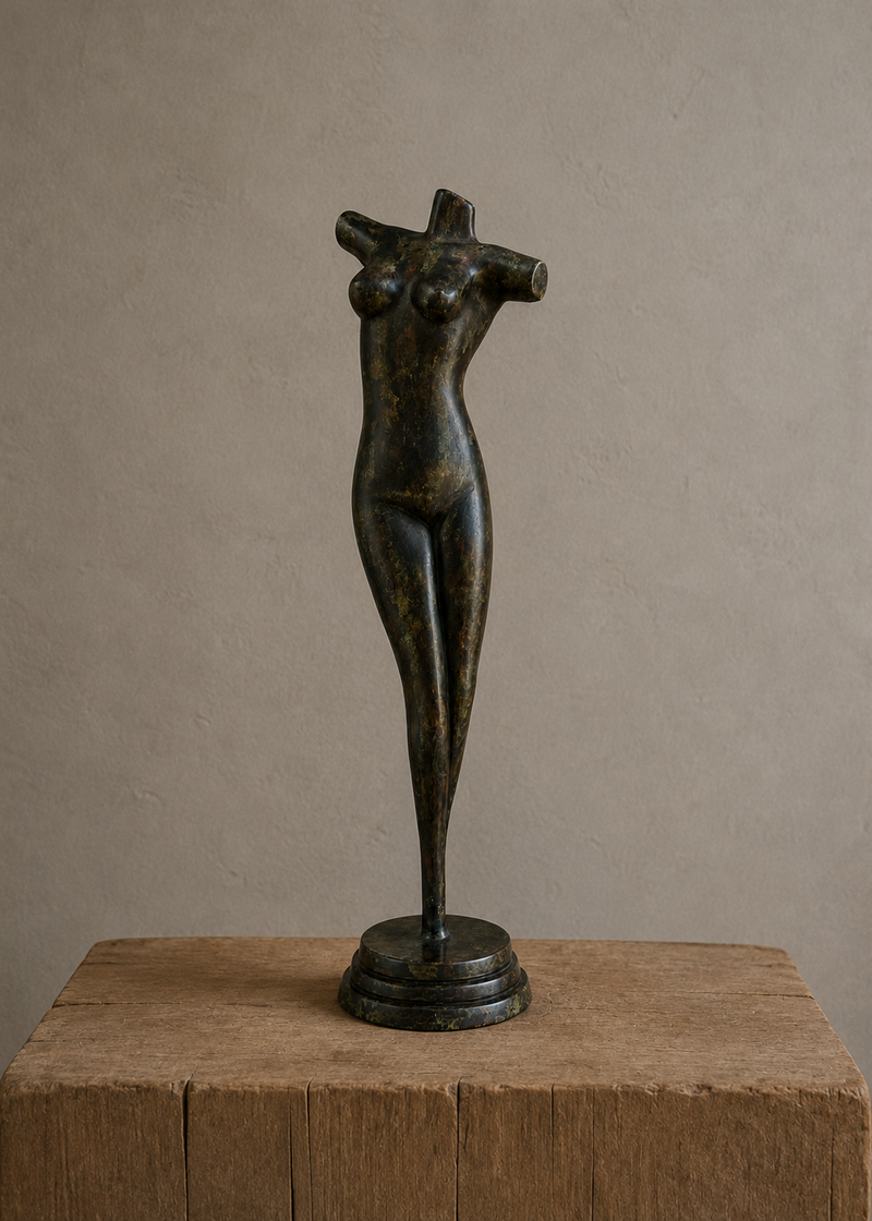 A dark bronze sculpture of a stylized female figure with crossed arms, displayed on a rustic wooden block against a neutral textured beige wall.
