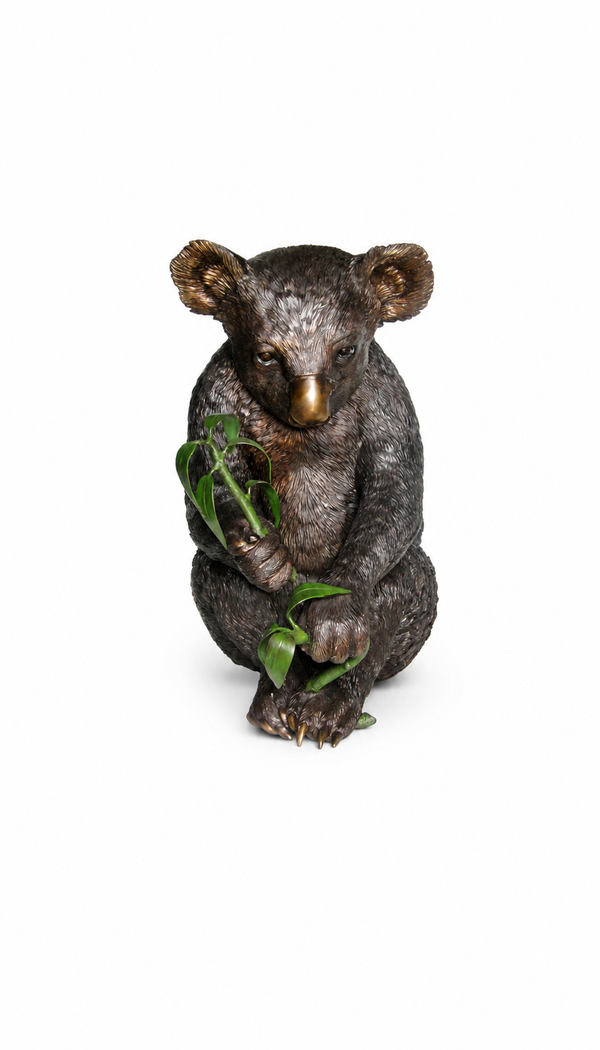 A koala bronze figurine posed sitting and holding green leaves, photographed against a plain white background.