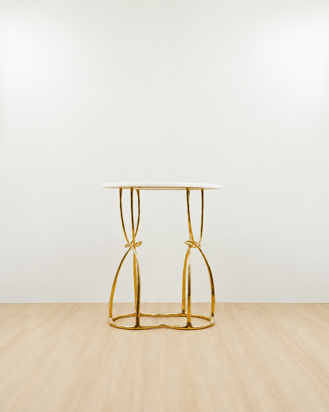 A modern console table with a white oval top and a polished gold brass base featuring twisted, knot-like legs, set against a minimalist white wall and light wood floor.