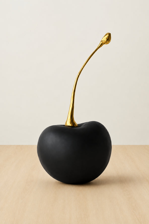 A decorative black cherry sculpture with a long, curved metallic gold stem, displayed on a light wood surface against a neutral off-white background.