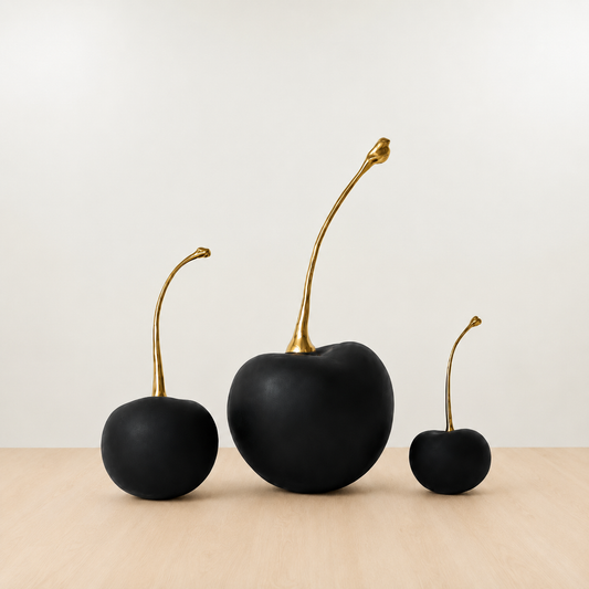Three matte black decorative fruit sculptures resembling cherries, each with a curved metallic gold stem, displayed on a light wood surface against a neutral off-white background.