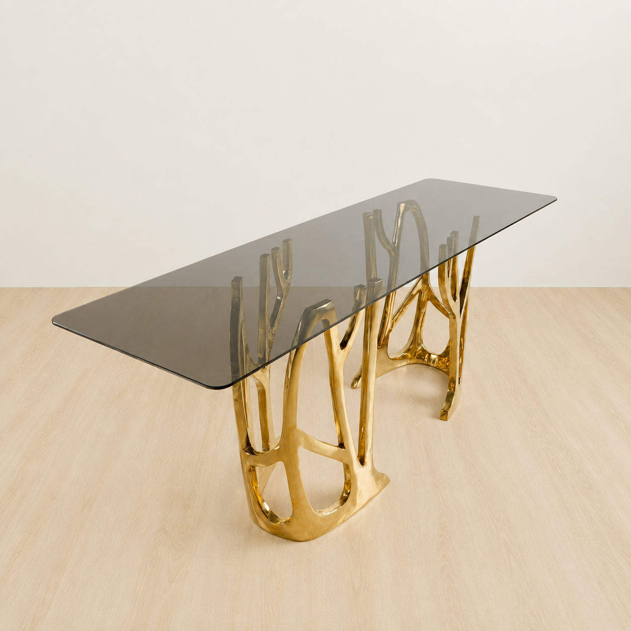 A modern rectangular console table with a dark smoked glass top and a sculptural, branching gold metal base, shown on a light wood floor against a neutral off-white wall.