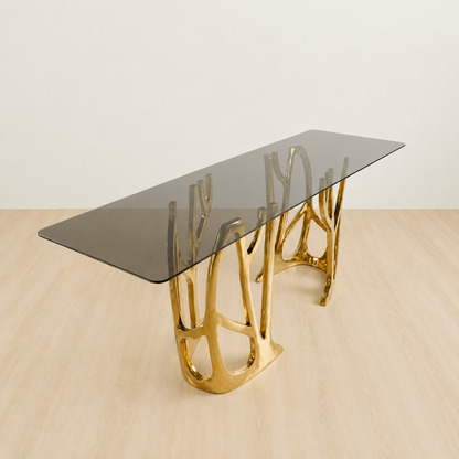 A modern rectangular console table with a dark smoked glass top and a sculptural, branching gold metal base, shown on a light wood floor against a neutral off-white wall.