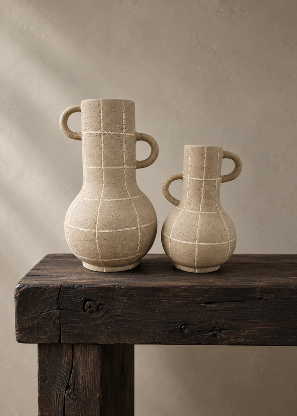 Two beige ceramic pitchers with loop handles sit on a dark rustic wooden table against a textured beige wall.