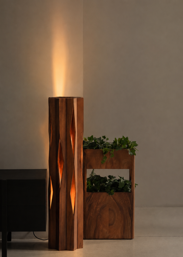 A modern wooden floor lamp with a tall, cylindrical body and carved diamond-shaped cutouts, glowing with warm light. Next to it is a matching wooden planter box with green plants, set against a minimalist beige wall and floor.