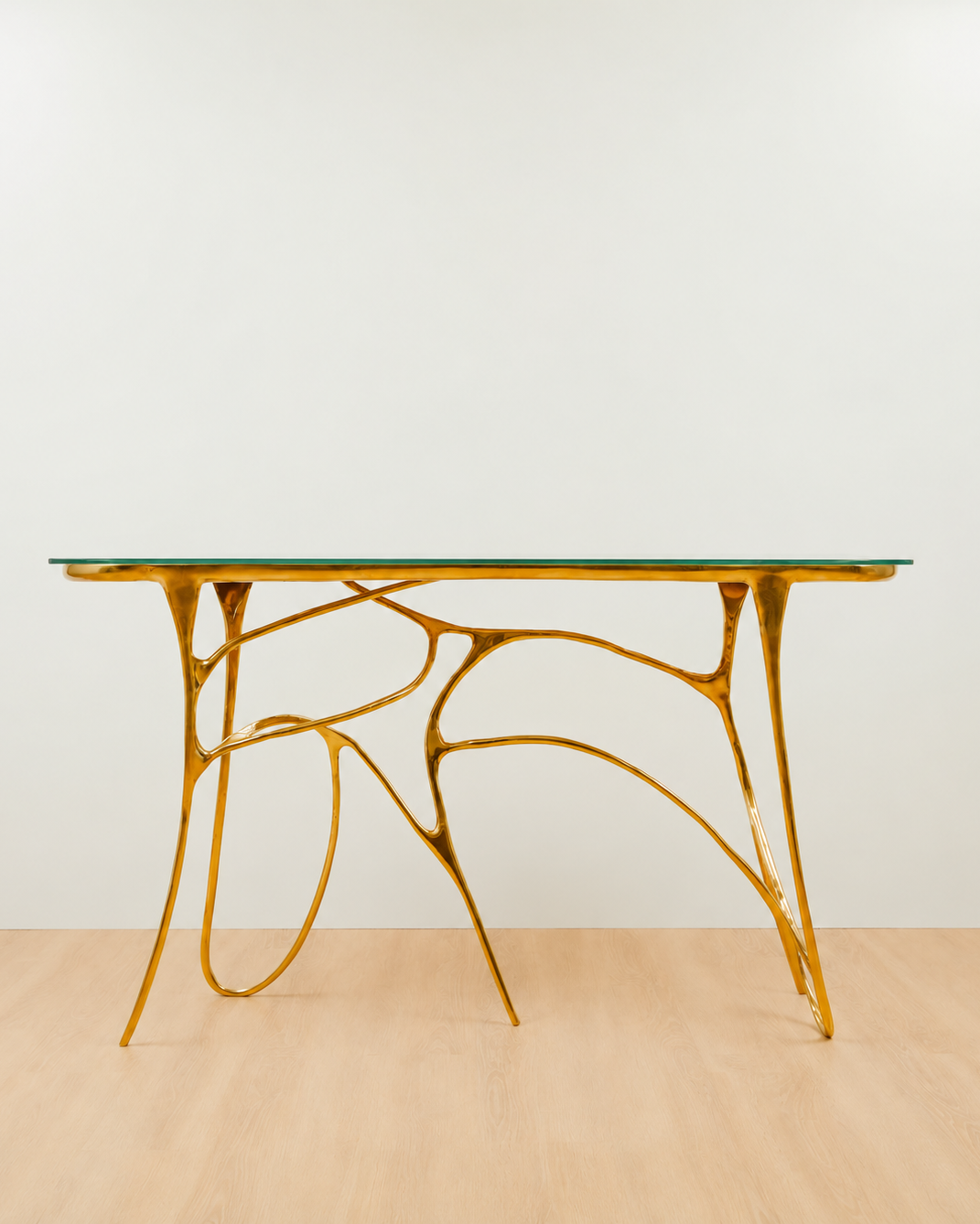 A modern console table with glass top and a sculptural, branching gold metal base, set against a plain white wall and light wood floor.