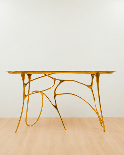 A modern console table with glass top and a sculptural, branching gold metal base, set against a plain white wall and light wood floor.