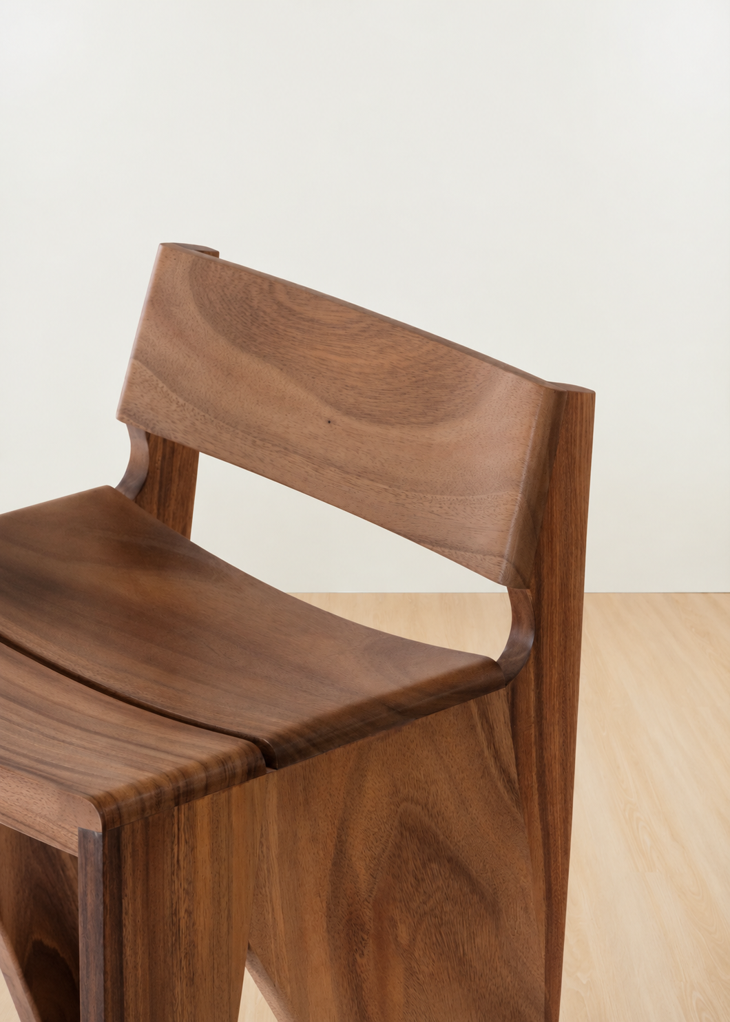 A close-up of a modern wooden chair with a rich walnut finish, showing the backrest and seat against a minimalist off-white wall and light wood floor.