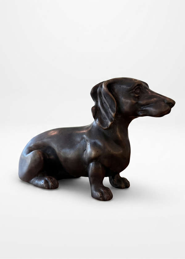 A dark bronze sculpture of a sitting dachshund dog, shown in profile against a clean, light gray studio background.