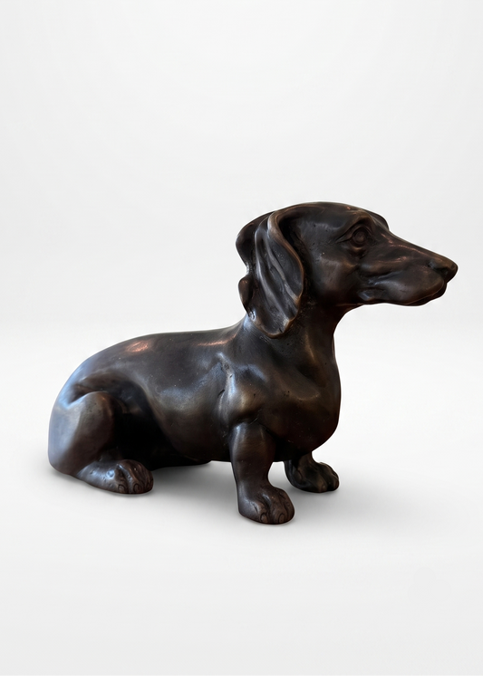 A dark bronze sculpture of a sitting dachshund dog, shown in profile against a clean, light gray studio background.