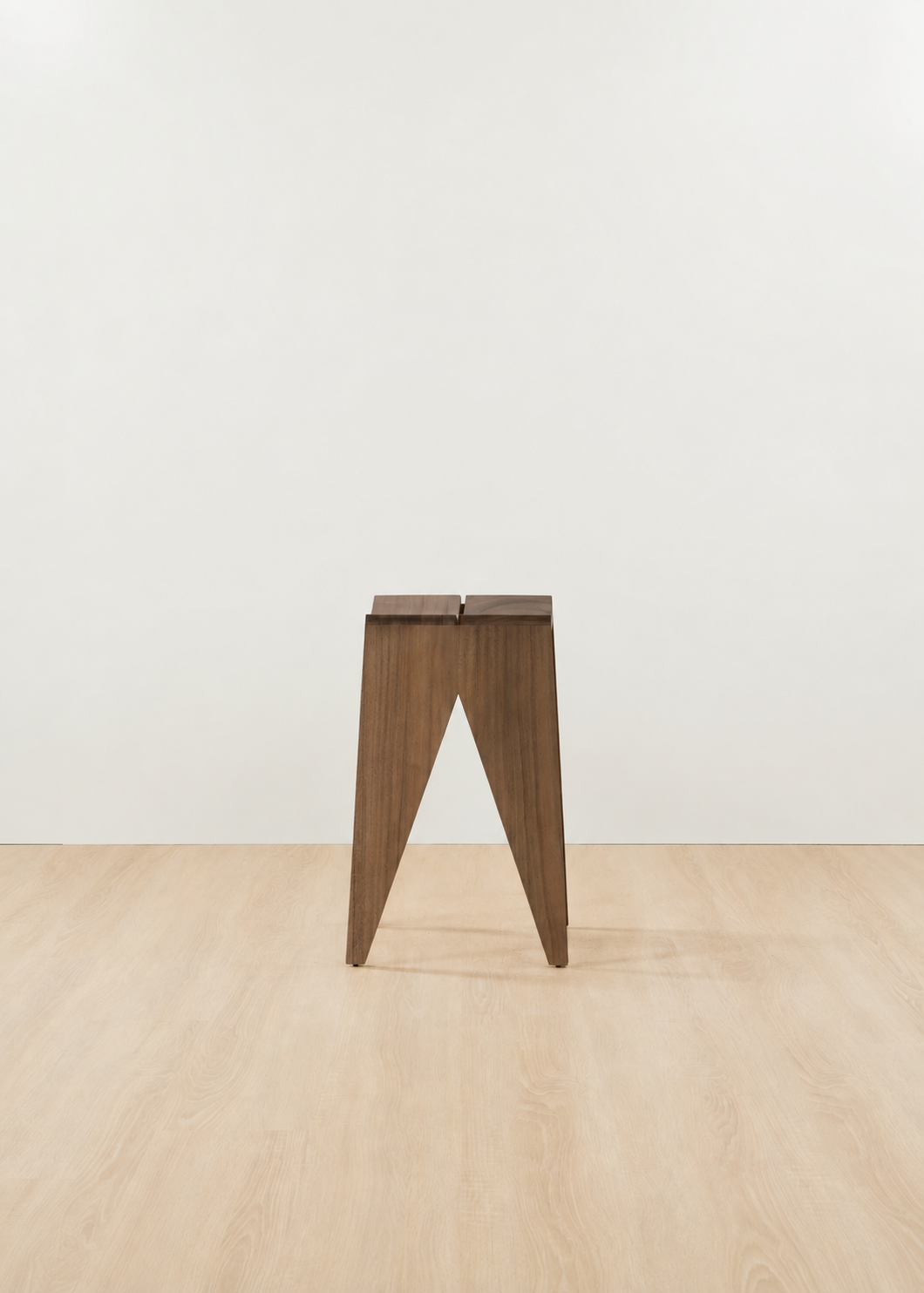 A minimalist wooden stool with a dark brown finish and a split rectangular seat, standing on a light wood floor against a plain white wall.