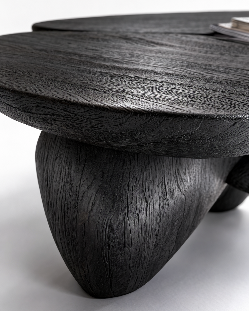 A close-up of a dark carved wooden table with a thick round top and a sculptural tree-like leg, showing detailed wood grain texture on a light gray background.