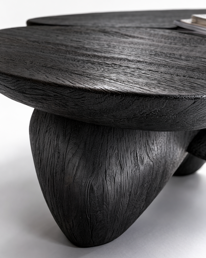 A close-up of a dark carved wooden table with a thick round top and a sculptural tree-like leg, showing detailed wood grain texture on a light gray background.