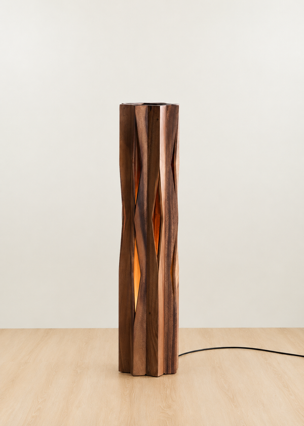 A modern sculptural floor lamp made of vertically stacked wavy wooden slats in warm brown and amber tones, illuminated from within, standing on a light wood floor against a plain off-white wall.
