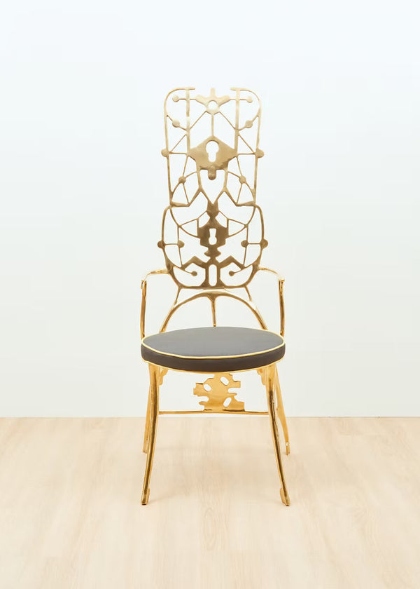 A decorative gold brass chair with an intricate openwork backrest and a dark gray upholstered seat, shown against a plain white wall and light wood floor.