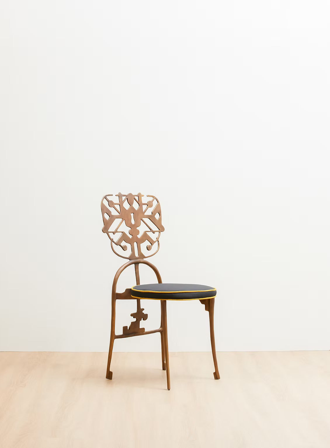 A vintage-style bras. chair with an ornate cutout backrest and a dark upholstered seat, set against a minimalist white wall and light wood floor.