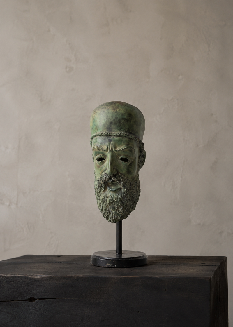 An antique-style green patina bronze sculpture of a bearded man's head wearing a hat, mounted on a black metal stand and displayed on a rustic dark wooden block against a textured beige plaster wall.