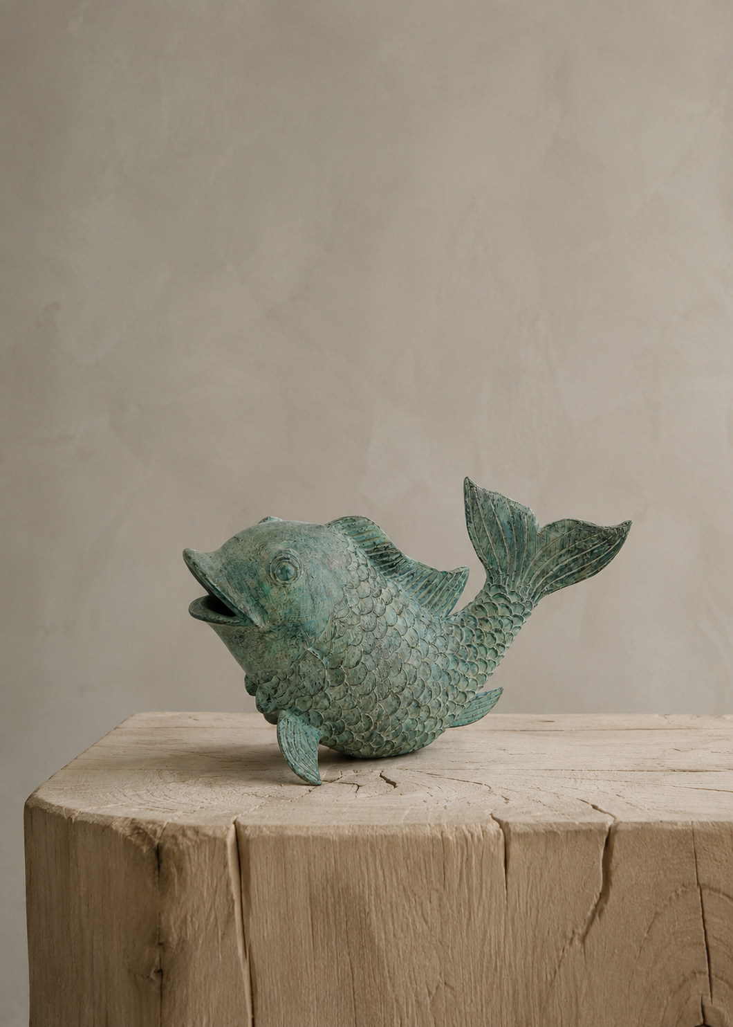 A small fish-shaped decorative bronze sculpture with a textured, weathered green patina finish, displayed on a rustic wooden block against a neutral beige plaster background.