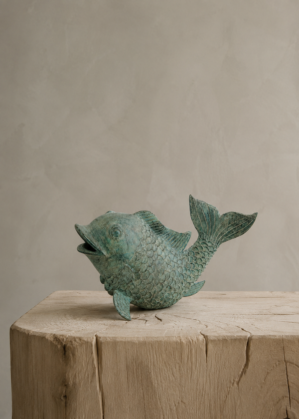 A small fish-shaped decorative bronze sculpture with a textured, weathered green patina finish, displayed on a rustic wooden block against a neutral beige plaster background.