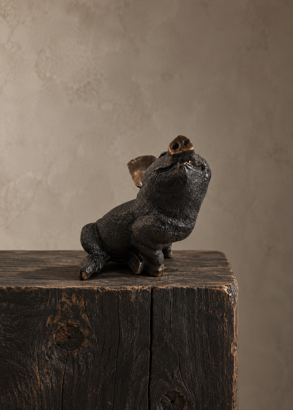 A small bronze sculptural figurine of a pig or piglet sitting on a rustic wooden block against a textured warm gray studio background.