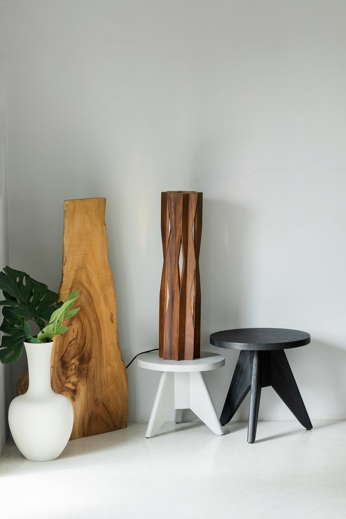 A styled interior scene with a tall sculptural wooden lamp or vase in the center, a black round side table to the right, a white pedestal supporting the lamp, a large natural wood slab leaning against the wall on the left, and a white vase with green leaves in the foreground.