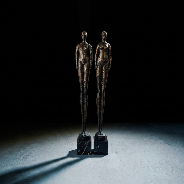 Two bronze nude human statues standing side by side on black bases, photographed in a dramatic studio setup with a dark background and spotlight on the floor.