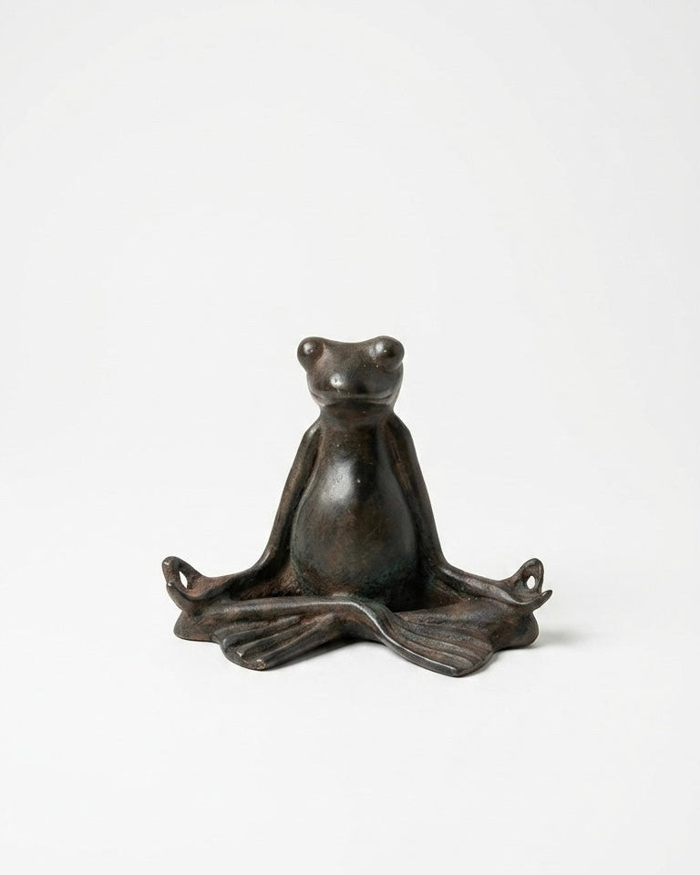 A small dark bronze frog figurine sitting in a meditative lotus pose, photographed against a seamless white studio background.