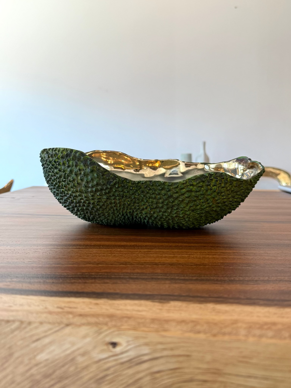 A decorative bowl shaped like a halved avocado, featuring a textured dark green exterior and a hammered metallic gold interior, resting on a wooden tabletop.