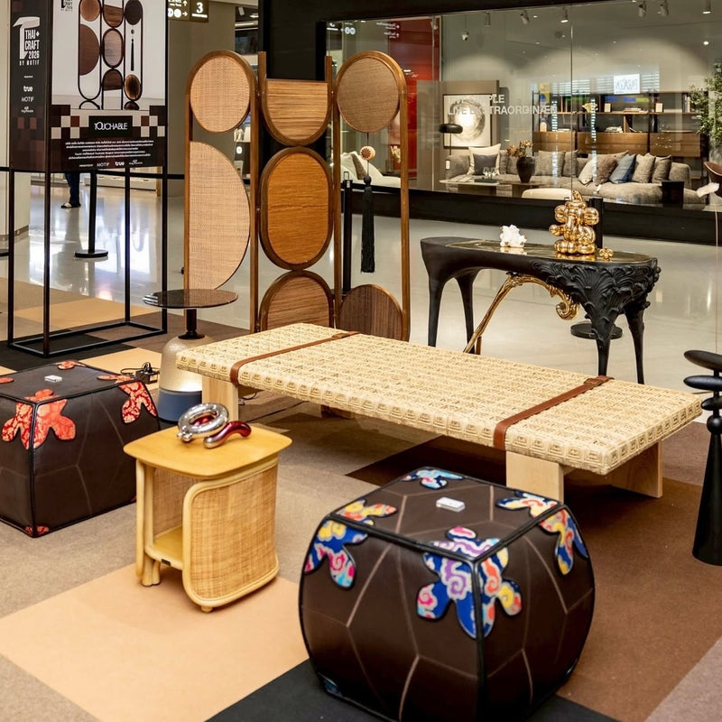 A styled furniture display in a showroom featuring a woven rattan daybed, a tall cane screen, a small wooden side table, and two dark brown ottomans with colorful floral patterns. The background shows a glass-walled retail space with other furniture pieces and decor.