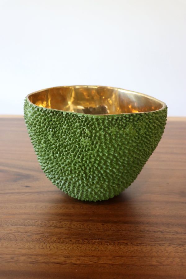 A textured green bowl with a shiny gold interior, displayed on a wooden table against a plain white background.