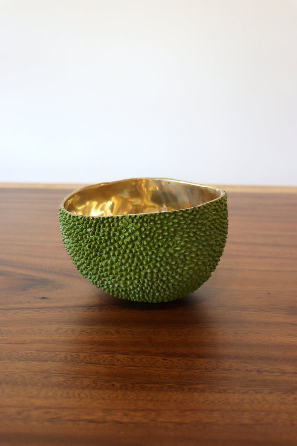 A textured green bowl with a hammered gold interior, resting on a dark wood tabletop against a plain white wall.