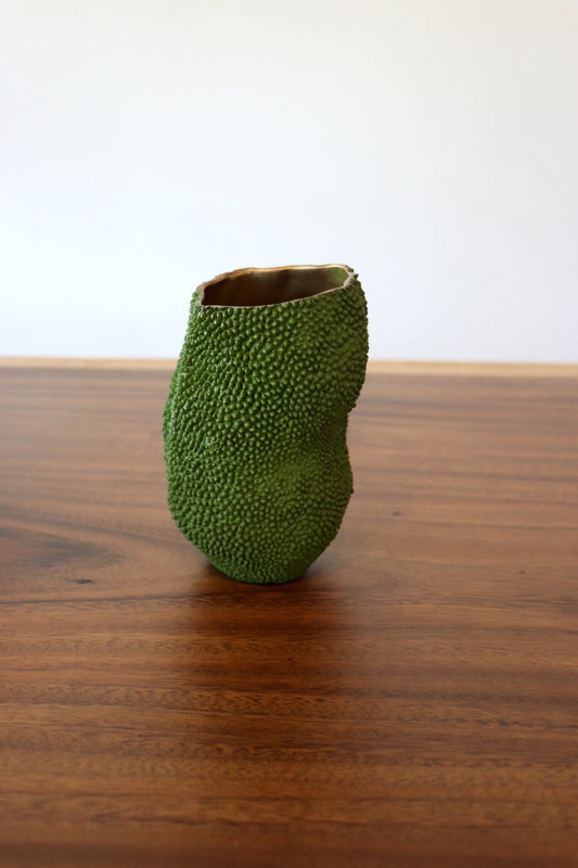A small sculptural vase made from a green textured fruit, standing on a polished wooden table against a plain white wall.