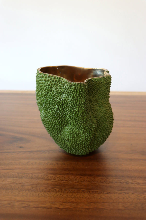 A green, textured, bumpy ceramic cup with a glossy brown interior, sitting on a wooden table against a plain white wall.
