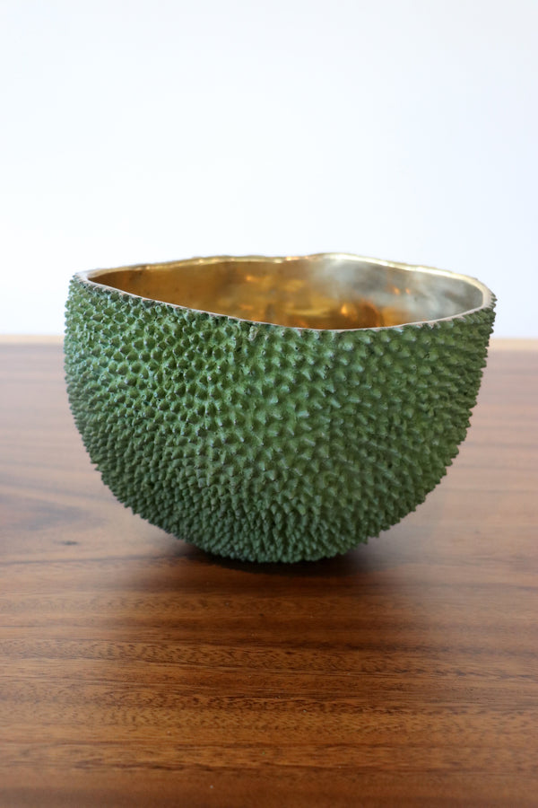A green textured bowl with a shiny gold interior, resting on a wooden tabletop against a white background.