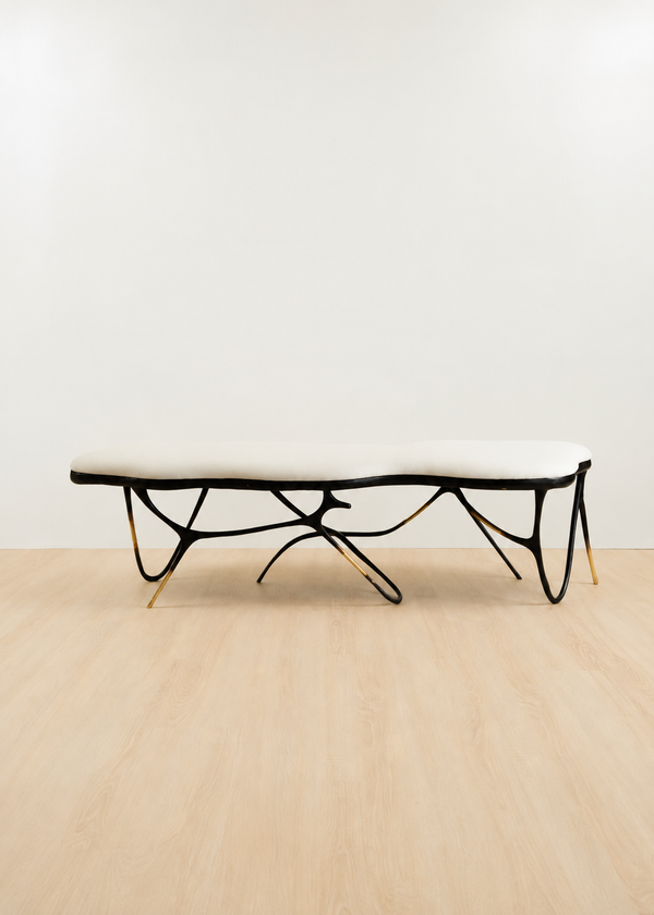 A modern bench with a white upholstered seat and a sculptural black brass frame with gold-toned legs, set against a minimalist white wall and light wood floor.