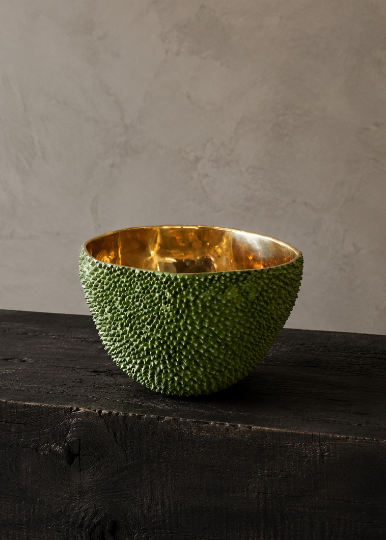 A decorative bowl with a textured green exterior resembling a rind and a hammered gold interior, displayed on a dark wooden block against a neutral gray wall.