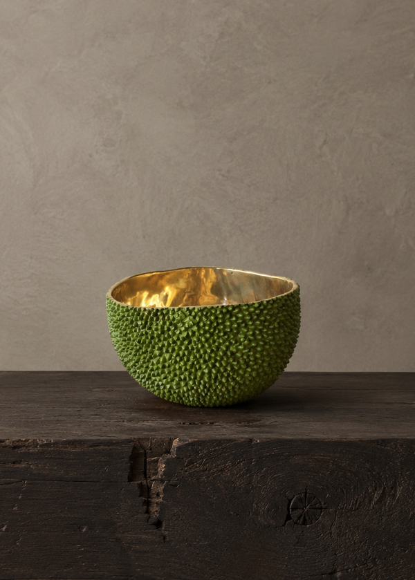 A green textured bowl with a shiny gold interior, styled on a dark rustic wooden surface against a neutral gray textured wall.