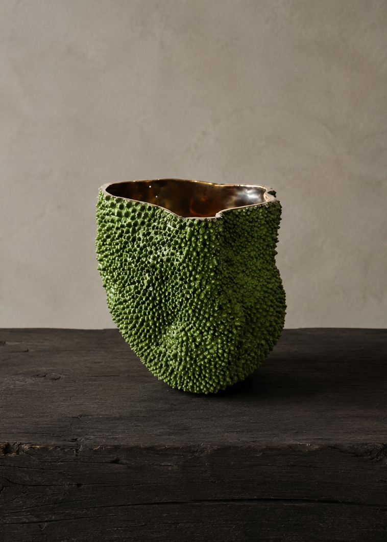 A textured green ceramic vase with a highly organic, bumpy surface and a glossy brown interior, displayed on a dark rustic wooden table against a neutral beige plaster wall.