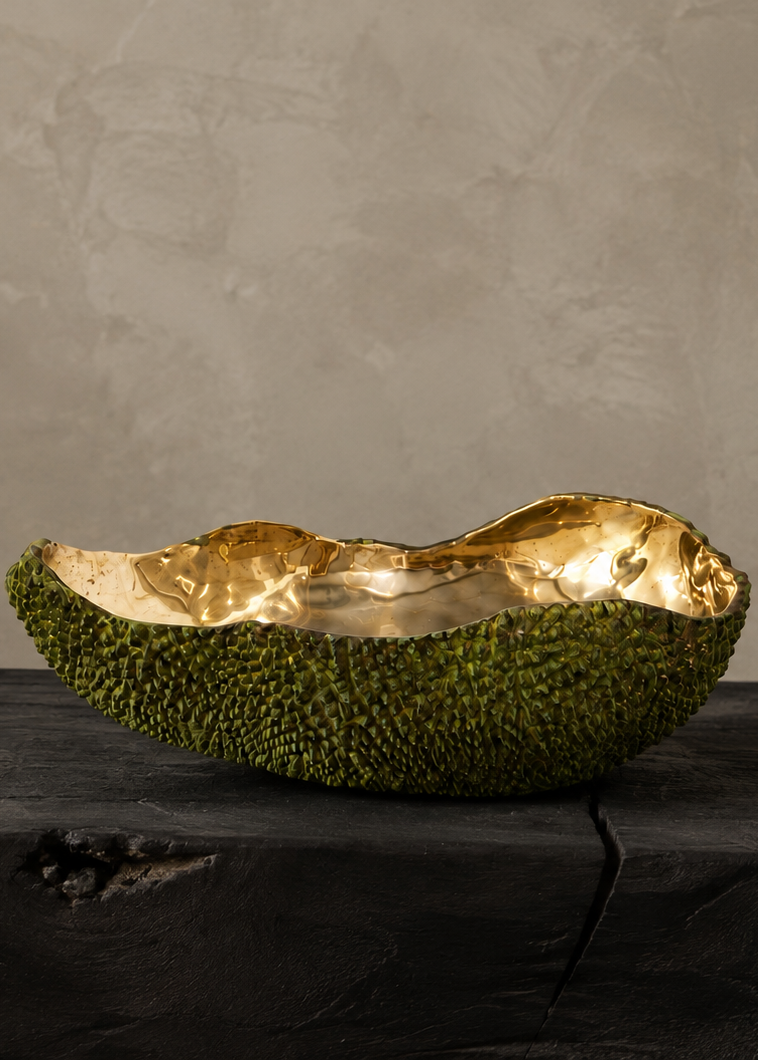 A decorative bowl crafted from a green jackfruit shell with a metallic gold leaf interior, displayed on a dark wooden block against a textured beige wall.