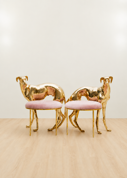 Two modern dog-shaped chairs with reflective gold brass bodies and round blush pink velvet seats, set against a minimalist off-white wall and light wood floor.